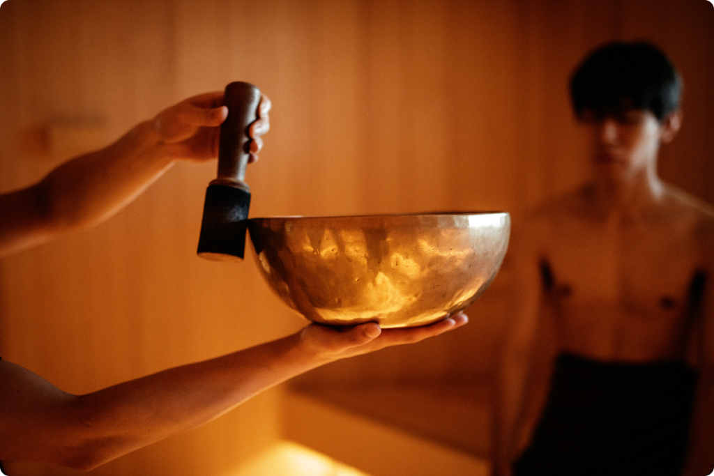 A singing bowl held in warm light inside a timber sauna, a guest resting behind